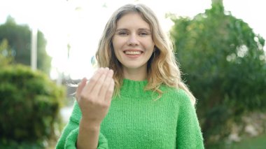 Young blonde woman smiling confident doing coming gesture with hand at park