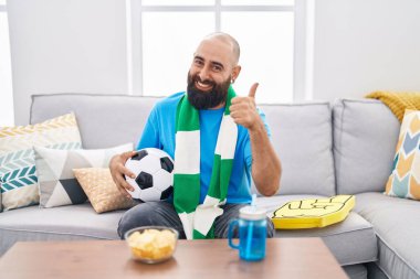 Young hispanic man with beard and tattoos football hooligan holding ball supporting team smiling happy and positive, thumb up doing excellent and approval sign 