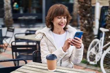 Middle age woman using smartphone sitting on table at coffee shop terrace