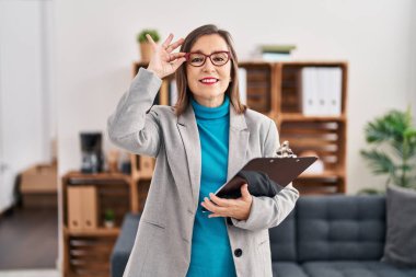 Middle age woman psychologist smiling confident holding clipboard at psychology center