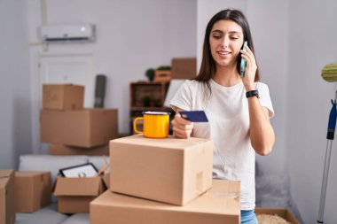 Young beautiful hispanic woman talking on smartphone holding credit card at new home
