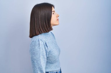 Mavi arka planda dikilen Latin kökenli genç kadın, yan tarafa bakıyor, rahat poz veriyor doğal bir yüz ve kendine güvenen bir gülümseme.. 