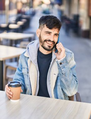 Young hispanic man talking on smartphone sitting on table at coffee shop terrace