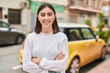 Genç İspanyol kadın gülümsüyor. Kollarını kavuşturup caddede duruyor.