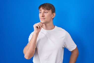Caucasian blond man standing over blue background with hand on chin thinking about question, pensive expression. smiling with thoughtful face. doubt concept. 