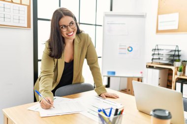 Young beautiful hispanic woman business worker writing on document standing at office