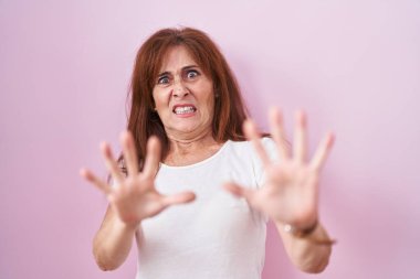 Middle age woman standing over pink background afraid and terrified with fear expression stop gesture with hands, shouting in shock. panic concept. 