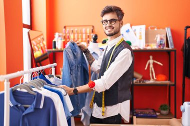Young hispanic man tailor smiling confident holding shirt of rack at atelier