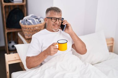 Orta yaşlı, kır saçlı, akıllı telefonda konuşan, yatak odasında kahve içen bir adam.