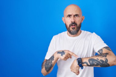 Hispanic man with tattoos standing over blue background in hurry pointing to watch time, impatience, upset and angry for deadline delay 