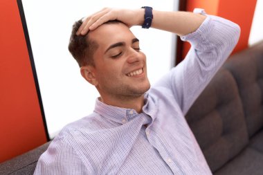 Young hispanic man relaxed with hand on head sitting on sofa at home