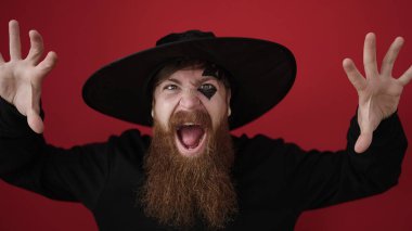 Young redhead man wearing wizard costume doing scare expression over isolated red background