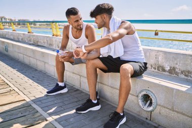 Two hispanic men sporty couple smiling confident looking watch sitting on bench at seaside