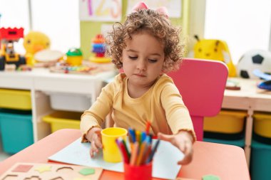 Adorable hispanic toddler student sitting on table drawing on paper at kindergarten