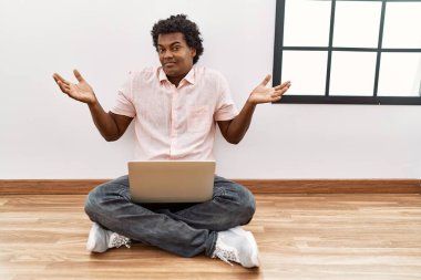 African man with curly hair using laptop sitting on the floor clueless and confused expression with arms and hands raised. doubt concept. 