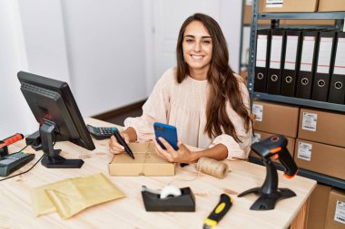 Young hispanic woman using smartphone writing on package order at storehouse