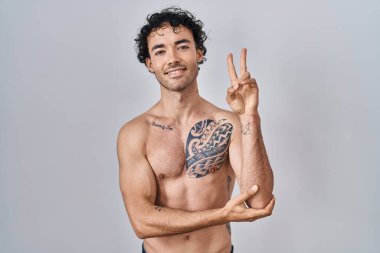 Hispanic man standing shirtless smiling with happy face winking at the camera doing victory sign. number two. 