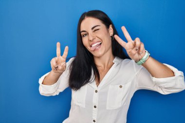 Young hispanic woman standing over blue background smiling with tongue out showing fingers of both hands doing victory sign. number two. 