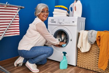 Çamaşır odasında orta yaşlı bir kadın çamaşır makinesini temizliyor.