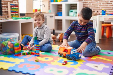 Anaokulunda yerde oturan iki çocuk inşaat bloklarıyla ve kamyon oyuncağıyla oynuyor.