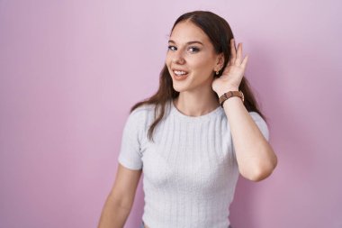 Young hispanic girl standing over pink background smiling with hand over ear listening an hearing to rumor or gossip. deafness concept. 