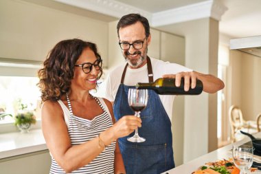 Orta yaşlı İspanyol çift mutfakta yemek pişiriyor ve bardaklara şarap döküyor.