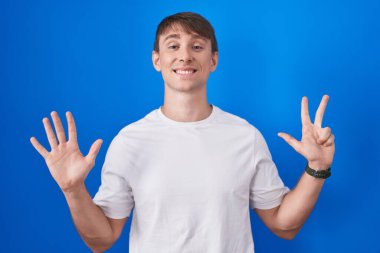 Caucasian blond man standing over blue background showing and pointing up with fingers number eight while smiling confident and happy. 