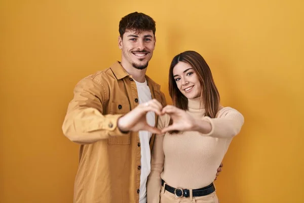 年轻的一对惊慌失措的情侣站在黄色的背景上 微笑着 用双手做着心形符号 浪漫的概念 — 图库照片