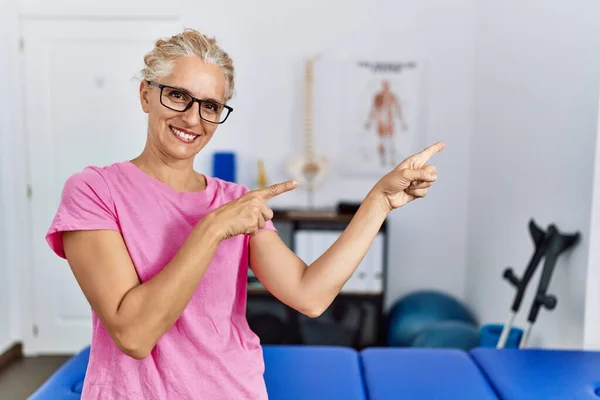 Middle age blonde woman at pain recovery clinic smiling and looking at the camera pointing with two hands and fingers to the side.