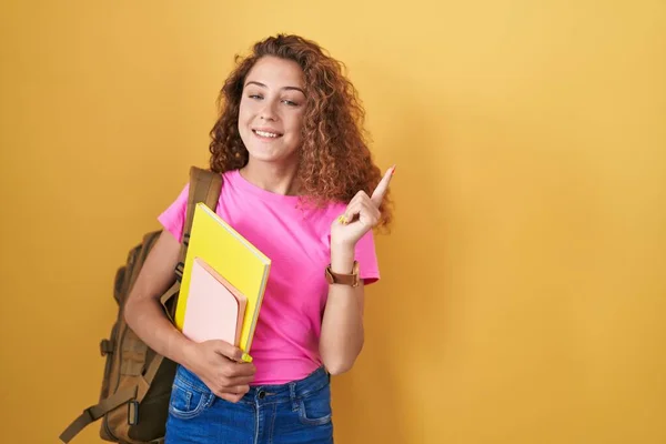 Young caucasian woman wearing student backpack and holding books cheerful with a smile on face pointing with hand and finger up to the side with happy and natural expression