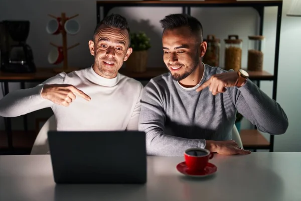 Homosexual couple using computer laptop looking confident with smile on face, pointing oneself with fingers proud and happy.