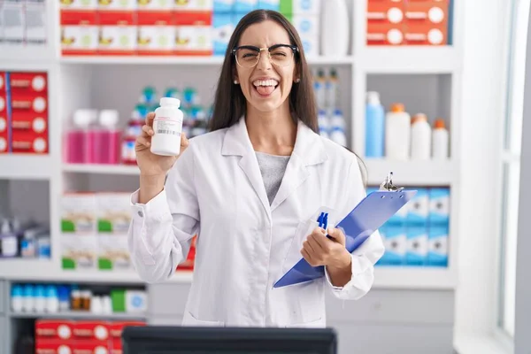 Young Brunette Woman Working Pharmacy Drugstore Holding Pills Sticking Tongue — Zdjęcie stockowe
