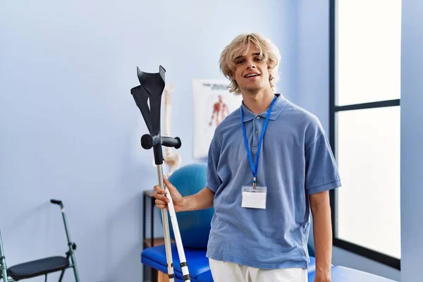 Young Blond Man Pysiotherapist Smiling Confident Holding Crutches Rehab Clinic — Stock Photo, Image