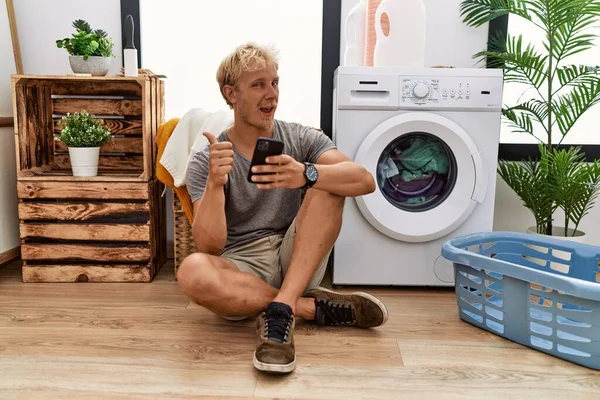 Young Blond Man Doing Laundry Using Smartphone Doing Happy Thumbs — Stok fotoğraf