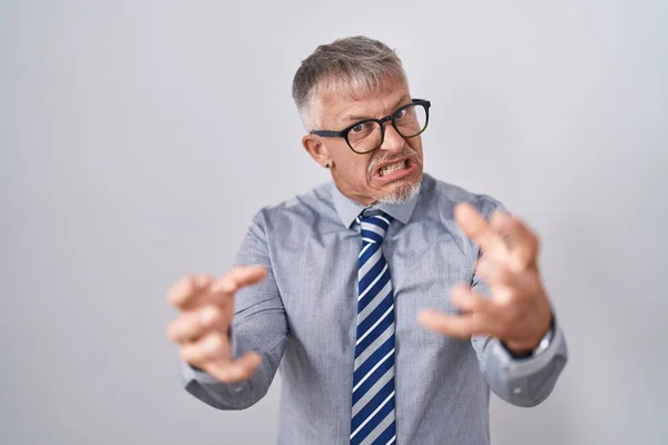 Hispanic business man with grey hair wearing glasses shouting frustrated with rage, hands trying to strangle, yelling mad