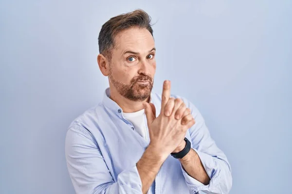 Middle age caucasian man standing over blue background holding symbolic gun with hand gesture, playing killing shooting weapons, angry face