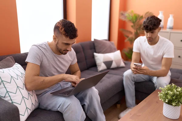 Two hispanic men couple using laptop and smartphone sitting on sofa at home