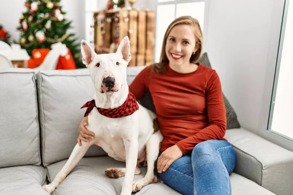 Young caucasian woman smiling confident hugging dog sitting by christmas tree at home