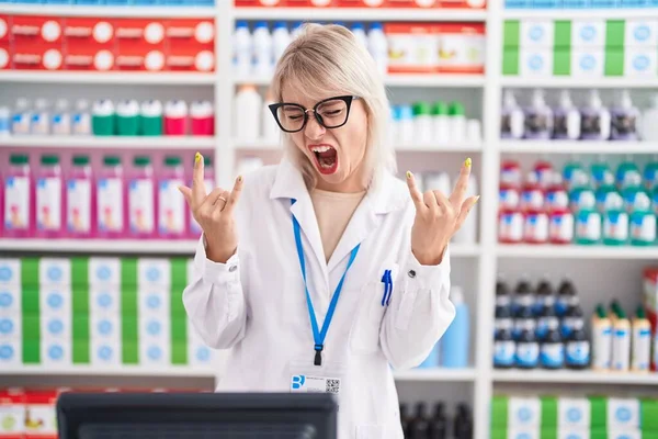 Young caucasian woman working at pharmacy drugstore shouting with crazy expression doing rock symbol with hands up. music star. heavy concept.