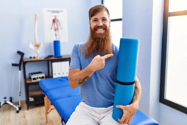 Redhead man with long beard holding yoga mat at rehabilitation clinic smiling happy pointing with hand and finger