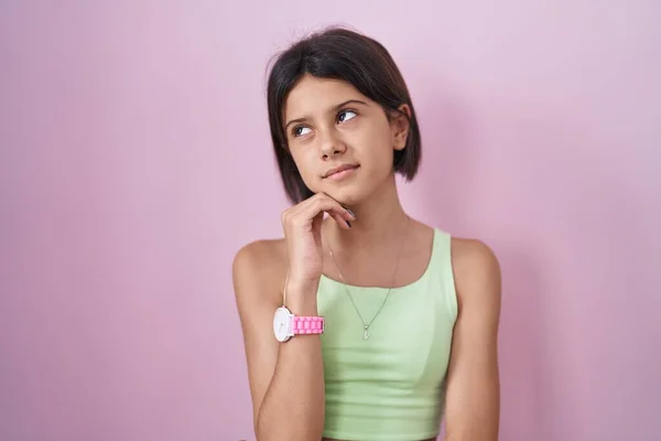 若い女の子のピンクの背景の上に手をあごの質問については 包括的な表現を考えて立っている 思慮深い顔で笑顔 疑わしい概念 — ストック写真