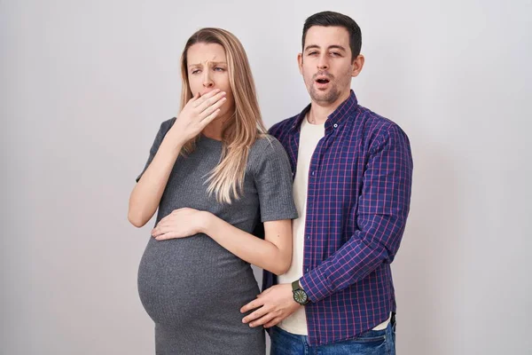 白い背景の上に立つ赤ちゃんを期待している若いカップルは 手で口をカバー疲れあくび退屈 落ち着きもなく眠気もなく — ストック写真