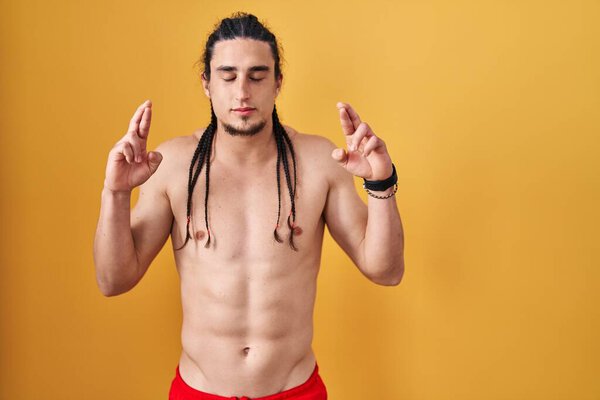 Латиноамериканец с длинными волосами, стоящий без рубашки на жёлтом фоне, жестом скрестив пальцы, улыбаясь с надеждой и закрыв глаза. удача и суеверие. 