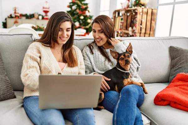 Two women using laptop sitting with dog by christmas tree at home