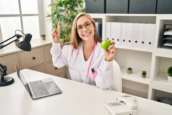 若いですブロンド医師女性ホールディング緑のりんごでザクリニック笑顔幸せ指して手と指へザ側 — ストック写真