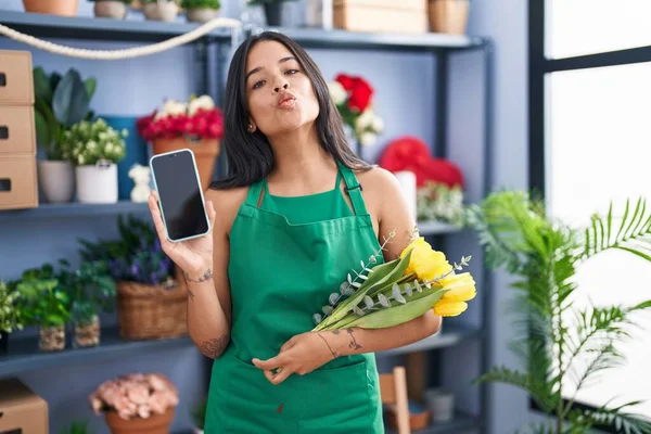 Brunette woman working at florist shop holding smartphone looking at the camera blowing a kiss being lovely and sexy. love expression.