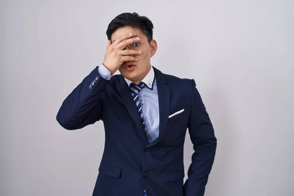 Young asian man wearing business suit and tie peeking in shock covering face and eyes with hand, looking through fingers with embarrassed expression.