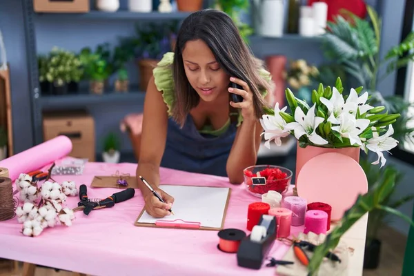 Young Hispanic Woman Florist Talking Smartphone Writing Document Florist Store — Zdjęcie stockowe