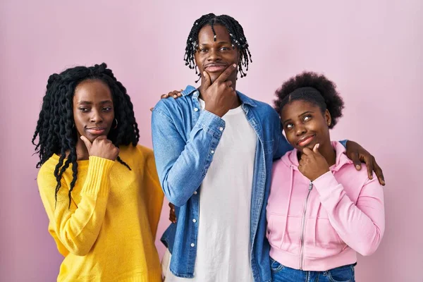 Group of three young black people standing together over pink background looking confident at the camera smiling with crossed arms and hand raised on chin. thinking positive.