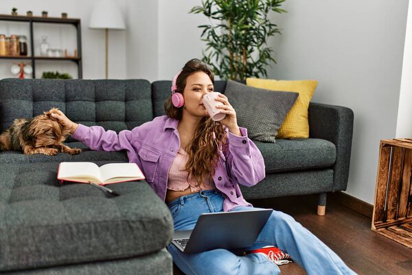 Молодая красивая латиноамериканка пьет кофе, прикасаясь к собаке, сидя дома на полу.